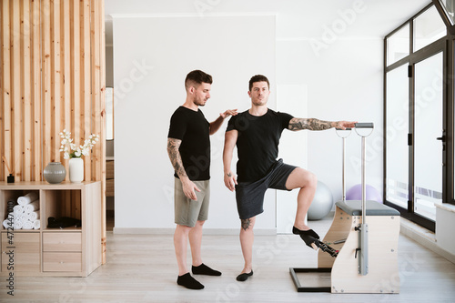 Man exercising at stability chair while male instructor supporting at pilates studio photo