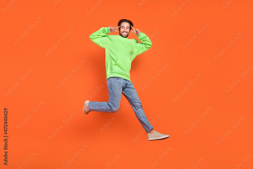 Full body photo of brunet millennial guy run show v-sign wear hoodie jeans footwear isolated on orange background