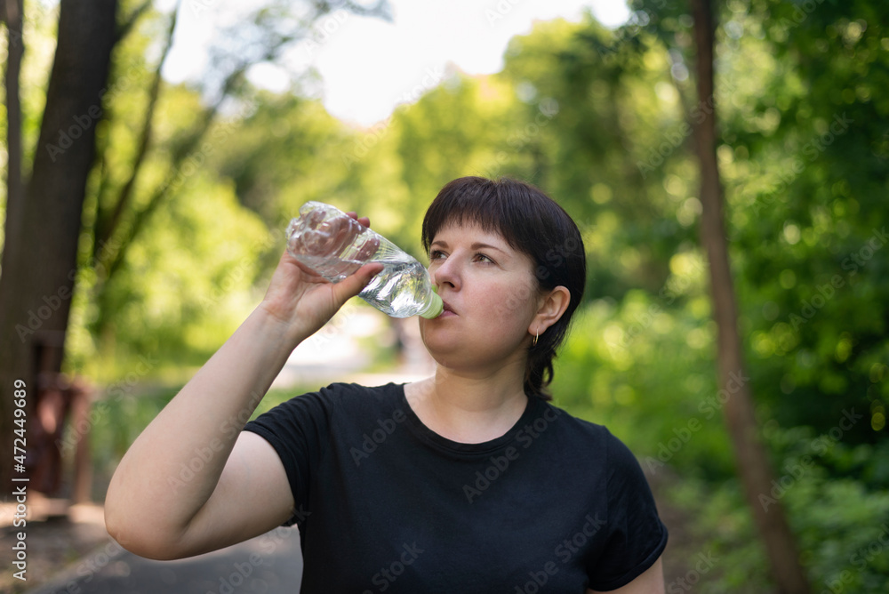 Young plus size woman drinks water after jogging in the summer park. Drinking regime during sports. Young plus size woman drinks water after jogging in the summer park. Drinking regime during sports.