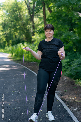 Plus size girl standing with a rope in his hands and looks at the camera in a summer park. Overweight young woman trains in the park. Vertical frame