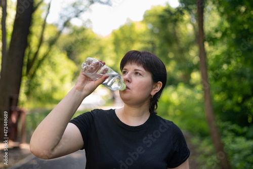 Young plus size woman drinks water after jogging in the summer park. Drinking regime during sports.