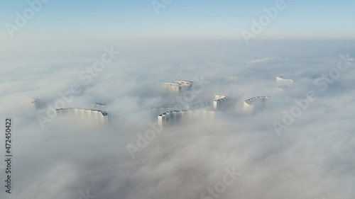 Silhouettes of tall houses through the incoming thick fog. Megalopolis in white clouds in the early morning at time.