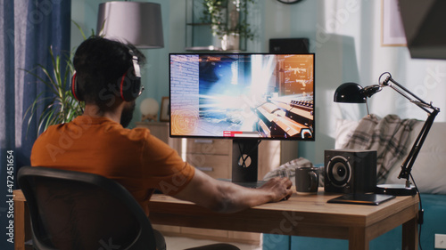 Side view of bearded Indian man in casual clothes and wireless headphones sitting at table and playing shooter on computer in weekend at home