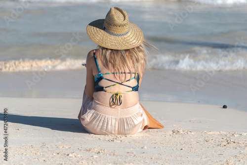 View from behind of a blonde girl with a hat seated on the beach looking at the sea on a sunny day.