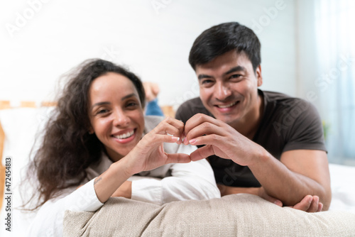 Happy Hispanic young couple making a heart shape with hands looking at the camera on the bed