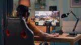 Back view of guy in casual clothes and headphones playing tank videogame on computer while sitting at desk in living room at home