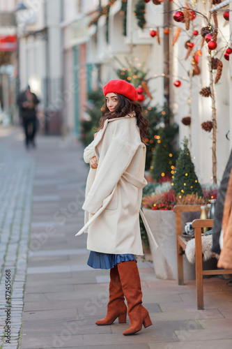 Christmas street style portrait of young beautiful woman in red beret walking in European city for winter holidays. Stylish model with curly hair is doing New Year's shopping.