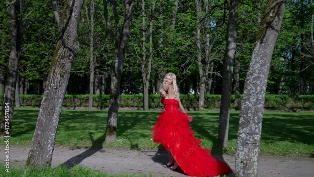 a blonde with big breasts and in a lush red dress runs along the path of the park between the trees. side view. the camera moves in parallel a blonde with big breasts and in a lush red dress runs along the path of the park between the trees. side view. the camera moves in parallel