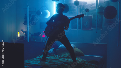 Pan around view of male teenager in glowing sunglasses playing loud music on electric guitar while standing on bed against blue neon lamps and he imagines that he is a rockstar on stage © Framestock