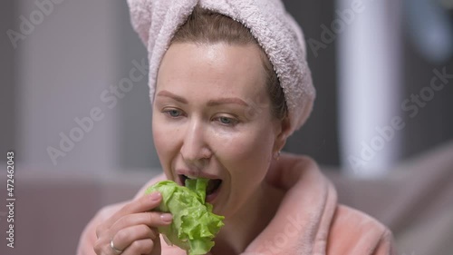 Tired dissatisfied young woman in hair towel eating green salad leaf sighing. Portrait of unhappy Caucasian millennial lady dieting at home in the morning. Femininity and self-discipline concept