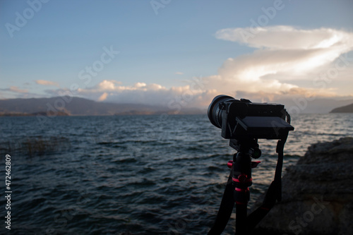 Close up to a modern camera over a tripod with blue lake, mountains and orange sunset scene at background