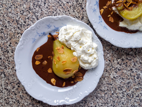 Top view of two plates with French dessert - poire belle Helene (poached caramelized pear with whipped cream, chocolate sauce and almond flakes).