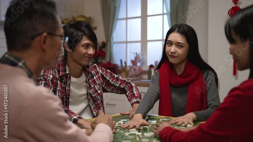 scheming asian son and daughter in red scarf smiling at other components while mixing mahjong tiles at table. indoor family recreation for lunar new year holiday concept