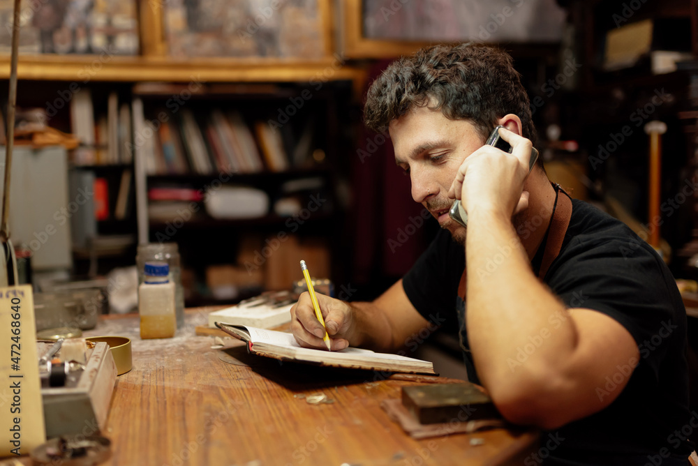 Young man in his craft workshop talking on the phone, picking up orders and writing down a notebook. Young man in his craft workshop talking on the phone, picking up orders and writing down a notebook.