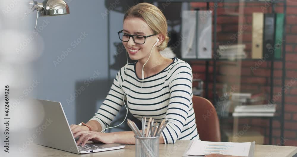 Beautiful blonde woman with glasses sitting in the office and typing on laptop enjoying her favorite music in headphones. Beautiful blonde woman with glasses sitting in the office and typing on laptop enjoying her favorite music in headphones.