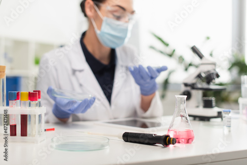 Electronic pipette, flask and petri dish near blurred scientist in laboratory.