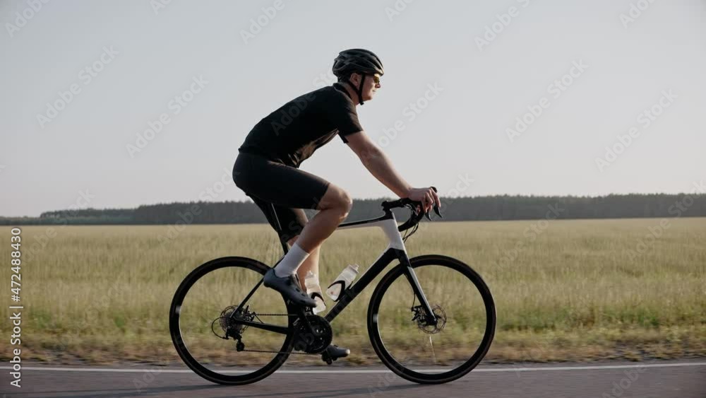 Man riding a road bike at the sunset on a highway near field, wearing helmet. Side shot of cyclist in black outfit riding road bicycle. 50 fps. 4k footage Man riding a road bike at the sunset on a highway near field, wearing helmet. Side shot of cyclist in black outfit riding road bicycle. 50 fps. 4k footage