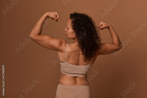 Rear view of a delighted confident middle aged African American woman in beige underwear looking at her arms and clenched fists, posing against colored background with copy space.