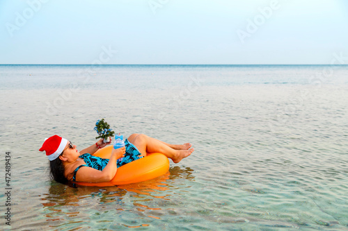 A woman in a swimming ring in a Santa Claus hat with a cocktail and a small Christmas tree in her hand near the shore in the sea. Shooting from the back.