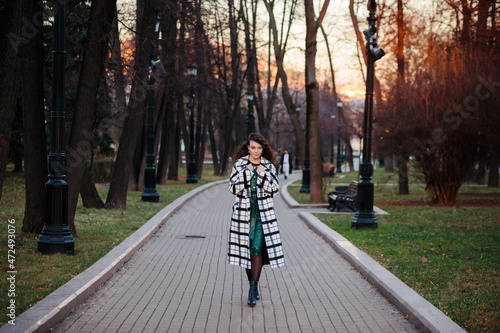 Fashionable woman in checkered elegant coat and green dress walking in park in autumn or in winter.