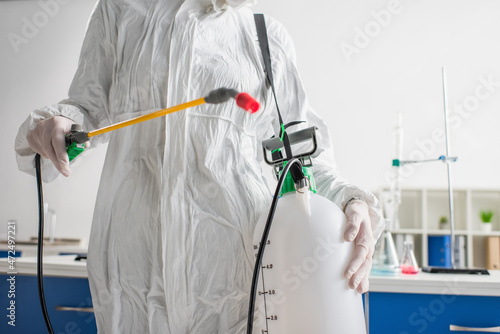 cropped view of scientist in protective suit holding sprayer while making disinfection of laboratory.