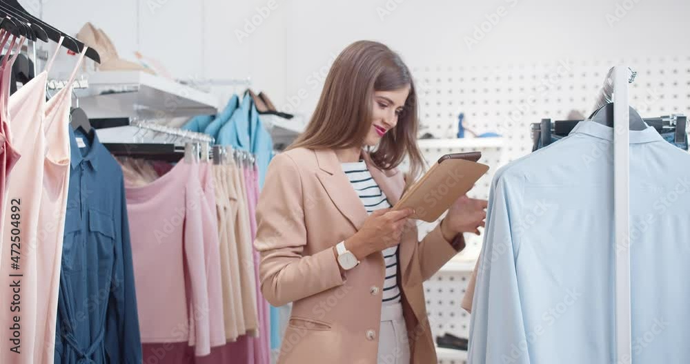 Young beautiful stylish woman in jacket standing in store holding tablet comparing prices and clothing size online. Young beautiful stylish woman in jacket standing in store holding tablet comparing prices and clothing size online.