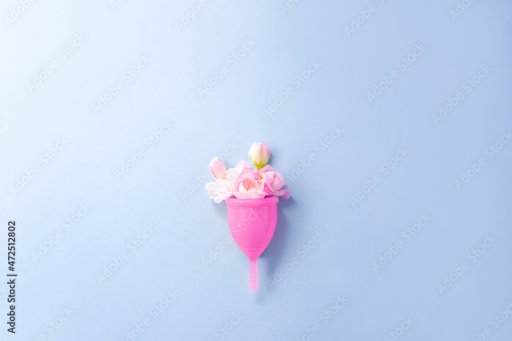 Menstrual cup and pink flowers on blue background. Concept of alternative methods of feminine hygiene. Copy space.