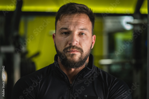 Front view close up portrait of young adult caucasian man with dark hair and beard in black gym suit looking to the camera in dark - Confident modern male dramatic portrait with copy space