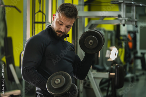 Side view portrait of young caucasian man male athlete bodybuilder training at the gym workout using dumbbells biceps curls wearing black shirt dark hair and beard standing weight lifting copy space