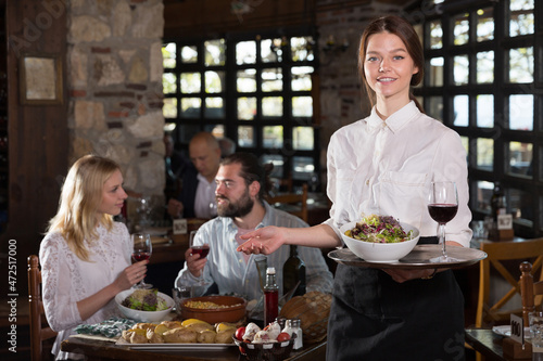 Portrait of smiling waitress of rustic restaurant holding tray with dishes  inviting guests for lunch