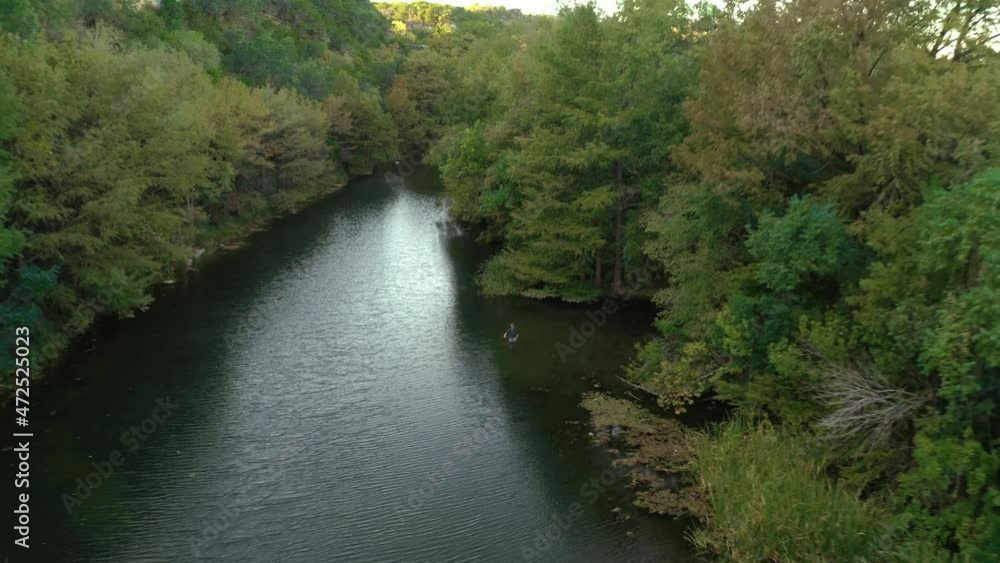 Flyfisherman in secluded river swimming hole casting fly rod while wading with lush tree scenery near shore, aerial drone flyaway to reveal forest shot in 4k Flyfisherman in secluded river swimming hole casting fly rod while wading with lush tree scenery near shore, aerial drone flyaway to reveal forest shot in 4k