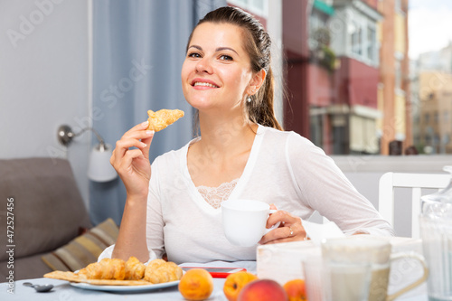 Portrait of young attractive woman with cup of tea eating croissant at table while breakfast