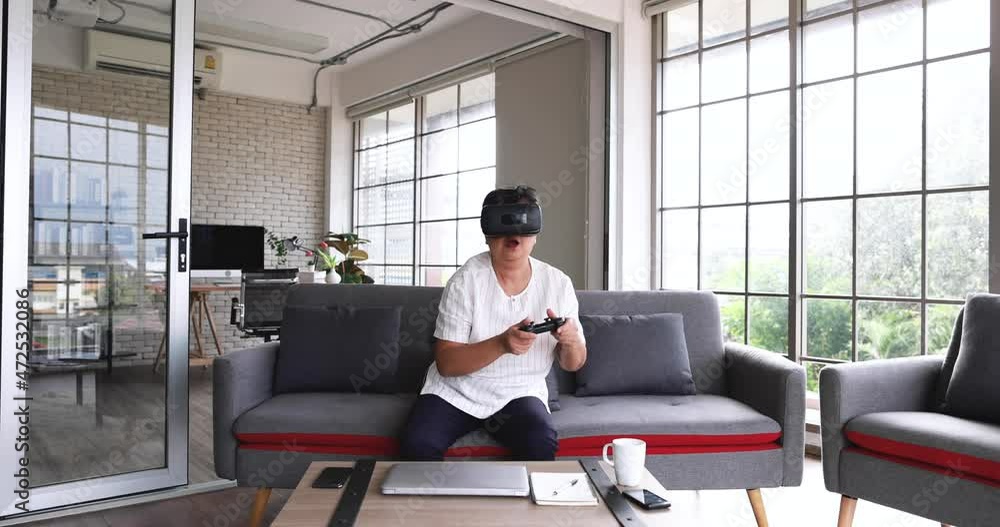 Asian old female sitting on sofa in living room wears vr virtual reality eyeglasses holding joystick while playing game with fun emotion. Concept of learning new technology for senior people. Asian old female sitting on sofa in living room wears vr virtual reality eyeglasses holding joystick while playing game with fun emotion. Concept of learning new technology for senior people.