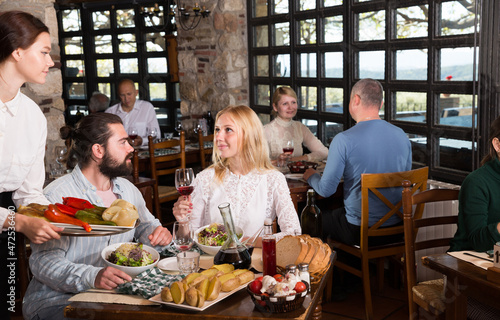 Young attractive waitress bringing delicious meals to visitors of cosy rustic style restaurant