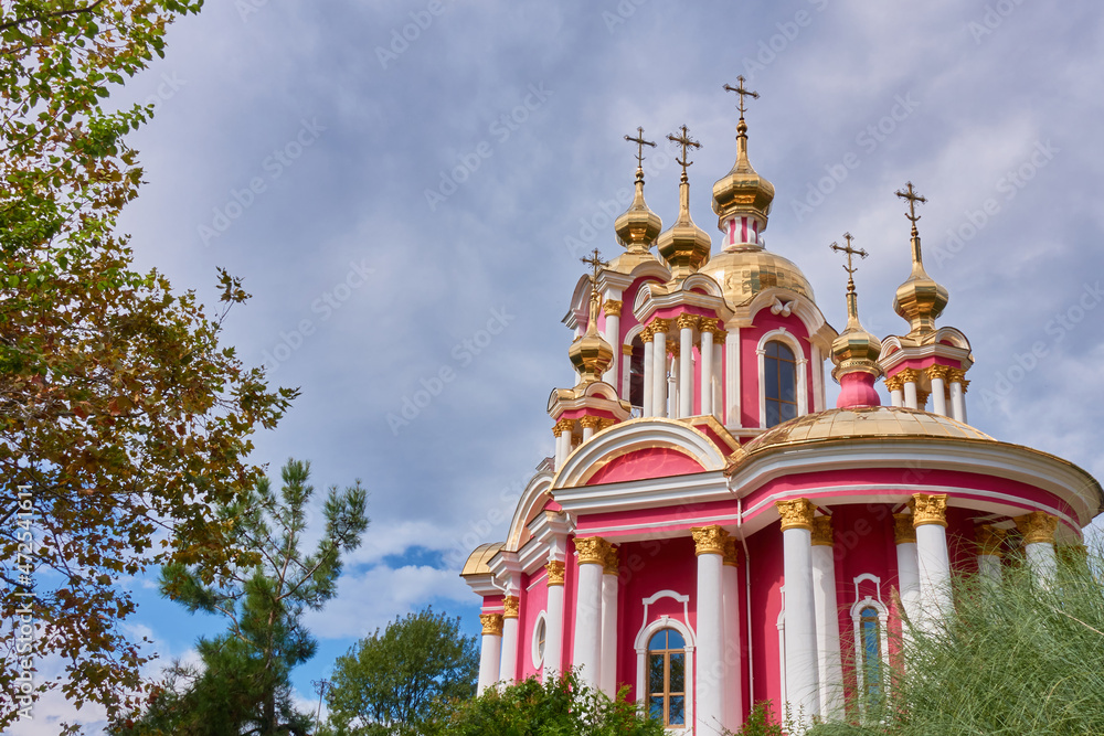 A red church with crosses, golden domes and white columns against a gray sky, surrounded by pines, trees and bushes.