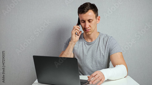 Valokuva Young brunette-haired man freelancer in grey t-shirt with broken forearm in gypsum band works distantly using laptop and talking via smartphone