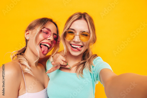 Two young beautiful smiling blond hipster female in trendy summer clothes. Sexy carefree women posing near yellow wall in studio. Positive models having fun. In sunglasses. Taking Pov photo selfie