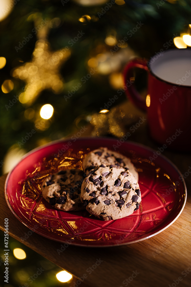 Cookies and warm milk left for Santa. Chocolate chip cookies and glass of milk in front of Christmas tree that is left blurred in background