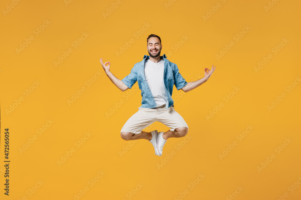 Full body young smiling fun happy man 20s wearing blue shirt jump high hold spreading hands in yoga om aum gesture relax meditate try to calm down isolated on plain yellow background studio portrait
