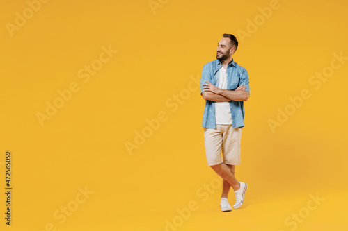 Full body young smiling happy fun minded cool man 20s wearing blue shirt white t-shirt hold hands crossed folded looking aside on copy space area isolated on plain yellow background studio portrait