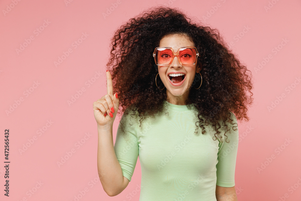 Insighted smart proactive young curly latin woman 20s years old wear mint t-shirt sunglasses holding index finger up with great new idea isolated on plain pastel light pink background studio portrait