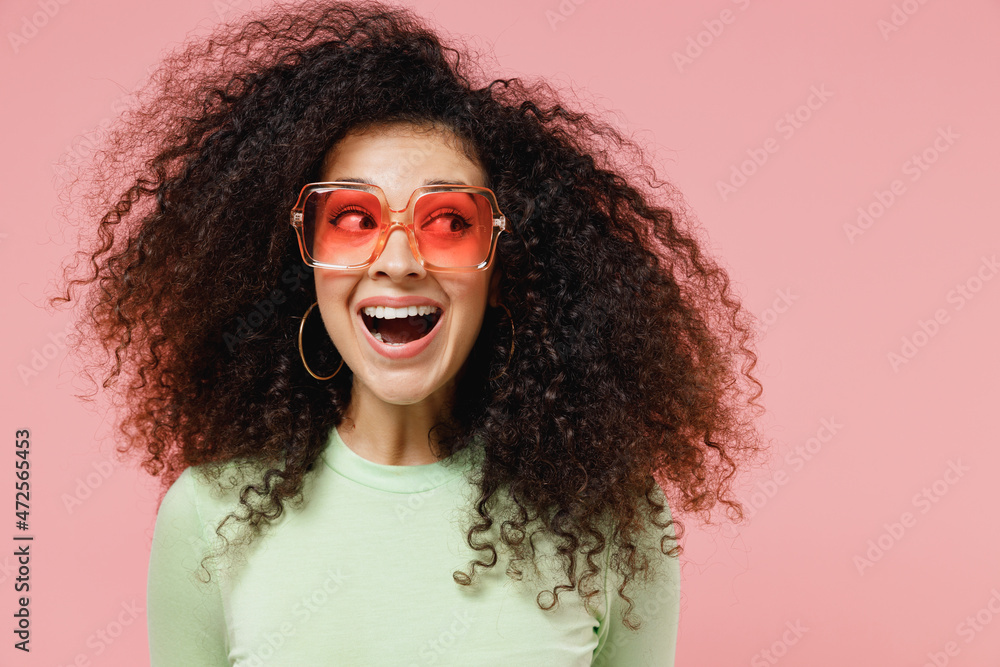 Close up fun joyful young curly latin woman 20s years old wears mint t-shirt sunglasses looking aside isolated on plain pastel light pink background studio portrait. People emotions lifestyle concept.