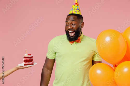 Young black gay man 20s in green t-shirt hat cone hold bunch of air inflated helium balloons celebrating birthday party, hand give cake with candle isolated on plain pastel pink background studio © ViDi Studio