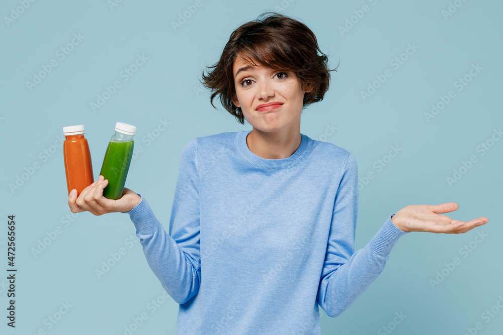 Young confused woman wear casual sweater hold pressed juice green orange vegetable smoothie as detox diet isolated on plain pastel light blue background studio portrait. People lifestyle food concept. Young confused woman wear casual sweater hold pressed juice green orange vegetable smoothie as detox diet isolated on plain pastel light blue background studio portrait. People lifestyle food concept.