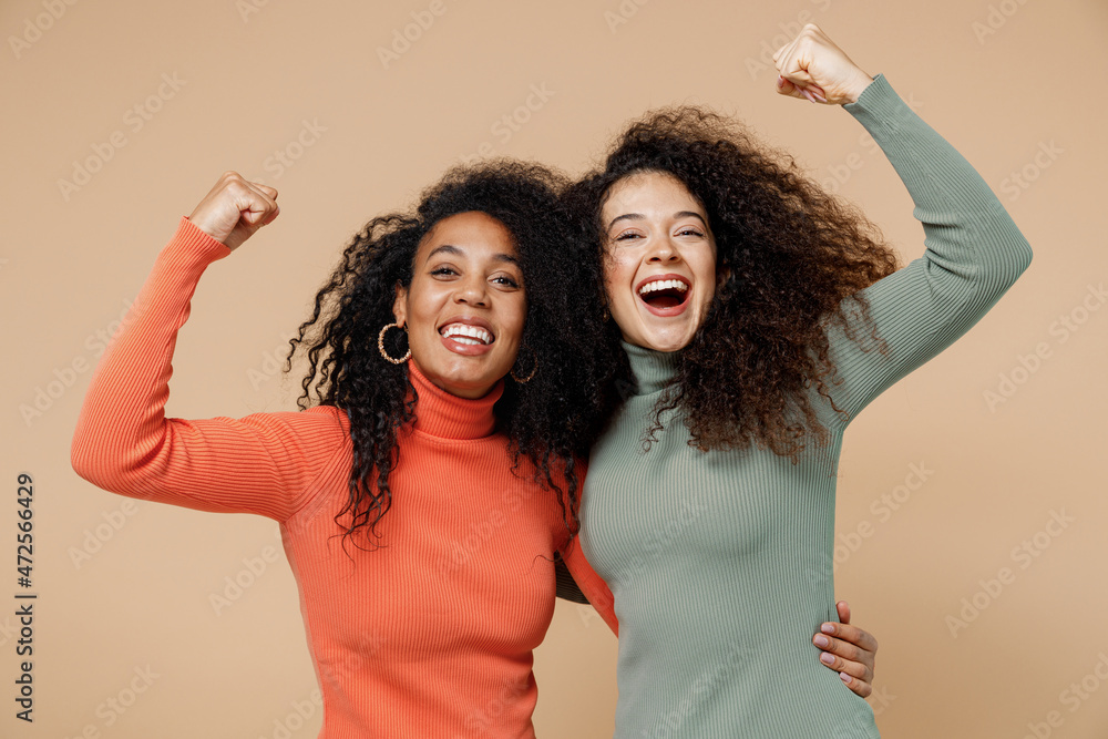 Two bright vivid young curly black women friends 20s wearing casual shirts clothes hug doing winner gesture celebrate clenching fists say yes isolated on plain pastel beige background studio portrait. Two bright vivid young curly black women friends 20s wearing casual shirts clothes hug doing winner gesture celebrate clenching fists say yes isolated on plain pastel beige background studio portrait.