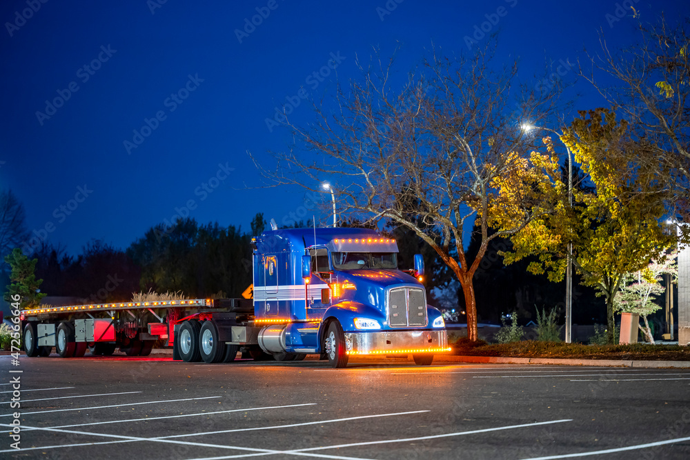 Blue classic big rig semi truck with turned on lights and flat bed semi