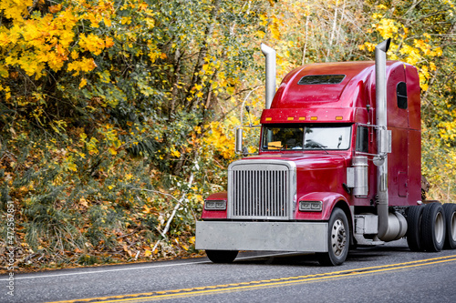 Burgundy big rig classic semi truck tractor running on the narrow autumn road to warehouse for the next loaded semi trailer