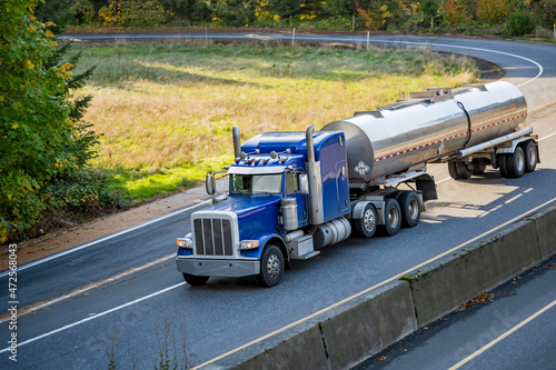 Classic blue bonnet big rig semi truck tractor transporting liquid cargo in tank semi trailer driving on the divided highway with round entrance road