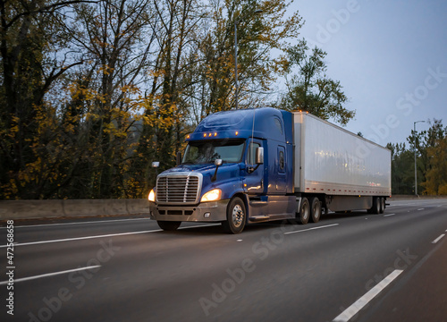 Dark blue big rig semi truck with turned on headlights transporting cargo in dry van semi trailer running on the evening twilight wide highway road with autumn trees on the side