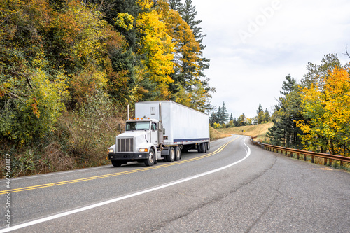 Day cab white big rig semi truck with dry van semi trailer transporting cargo driving on the winding narrow road through the autumn forest in Columbia Gorge area